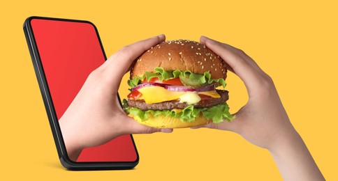 Image of Food ordering and delivery service. Woman eating burger with one hand sticking out of mobile phone display against orange background, closeup