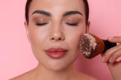 Photo of Beautiful woman applying makeup with brush on pink background, closeup