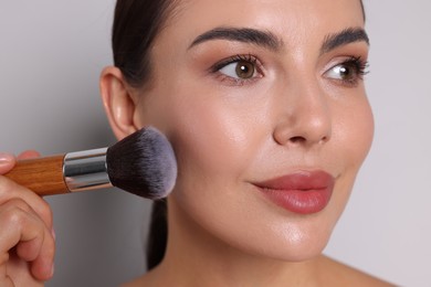 Photo of Beautiful woman applying makeup with brush on light grey background, closeup