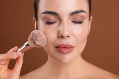 Photo of Beautiful woman applying makeup with brush on brown background, closeup