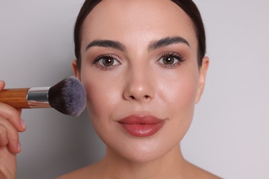 Photo of Beautiful woman applying makeup with brush on light grey background, closeup