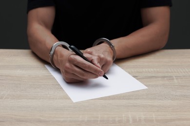 Photo of Man in metal handcuffs writing something at wooden table against dark background, closeup