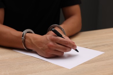 Photo of Man in metal handcuffs writing something at wooden table against dark background, closeup