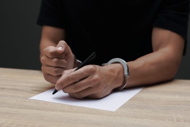 Photo of Man in metal handcuffs writing something at wooden table against dark background, closeup