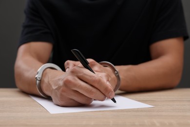 Photo of Man in metal handcuffs writing something at wooden table against dark background, closeup