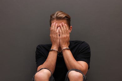 Photo of Man in metal handcuffs covering face with hands near grey wall