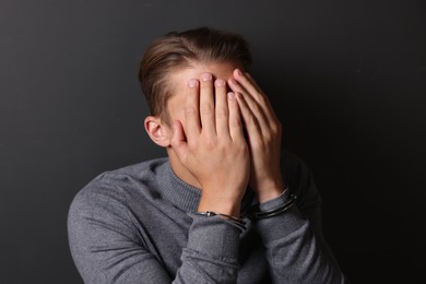 Photo of Man in metal handcuffs covering face with hands near grey wall