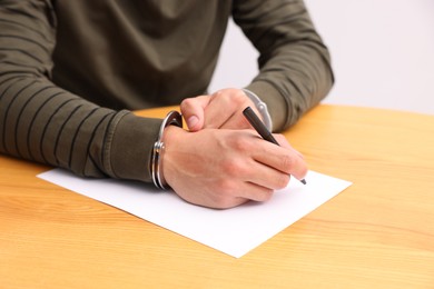 Photo of Man in metal handcuffs writing something at wooden table against light background, closeup