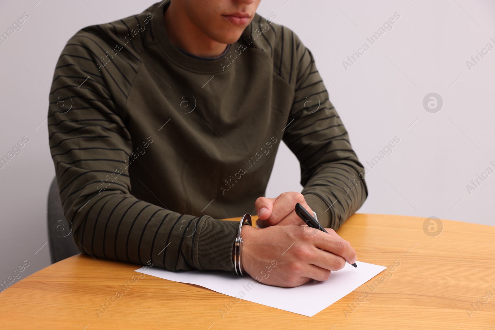 Man in metal handcuffs writing something at wooden table against light grey background, closeup Photo of Man in metal handcuffs writing something at wooden table against light grey background, closeup