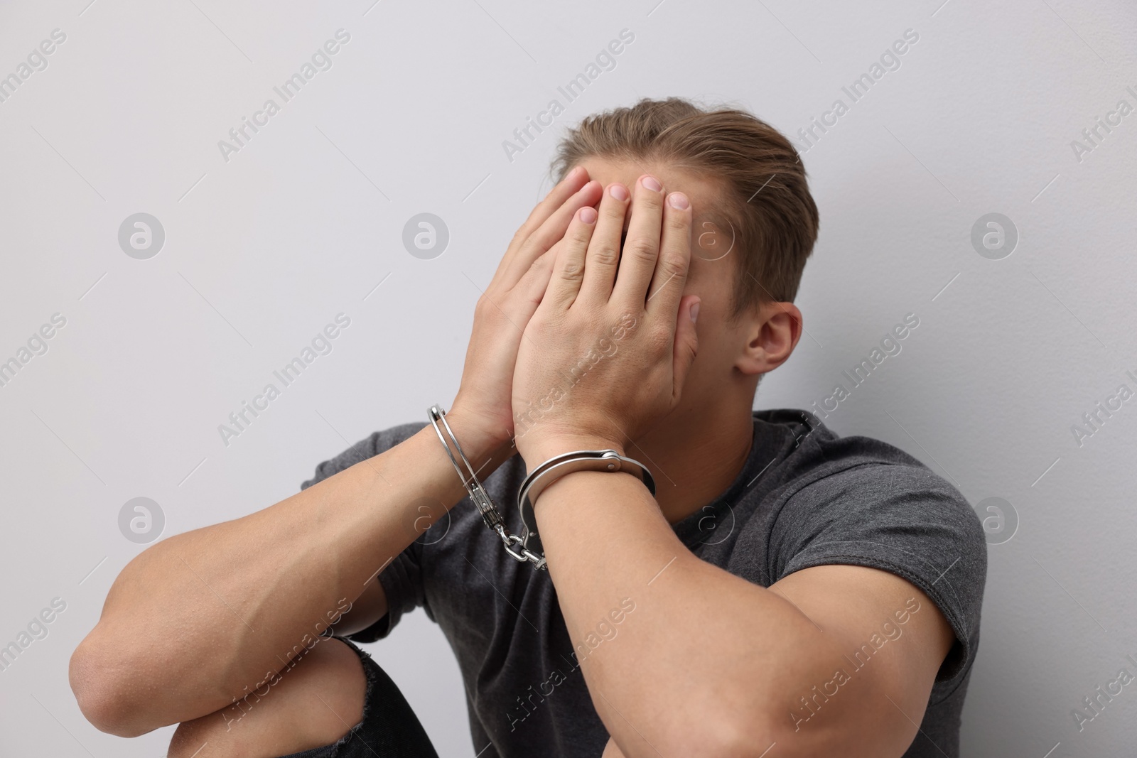 Man in metal handcuffs covering face with hands near light grey wall Photo of Man in metal handcuffs covering face with hands near light grey wall