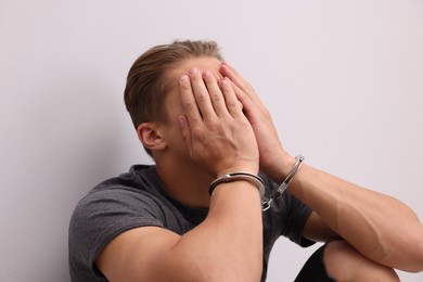 Photo of Man in metal handcuffs covering face with hands near light grey wall