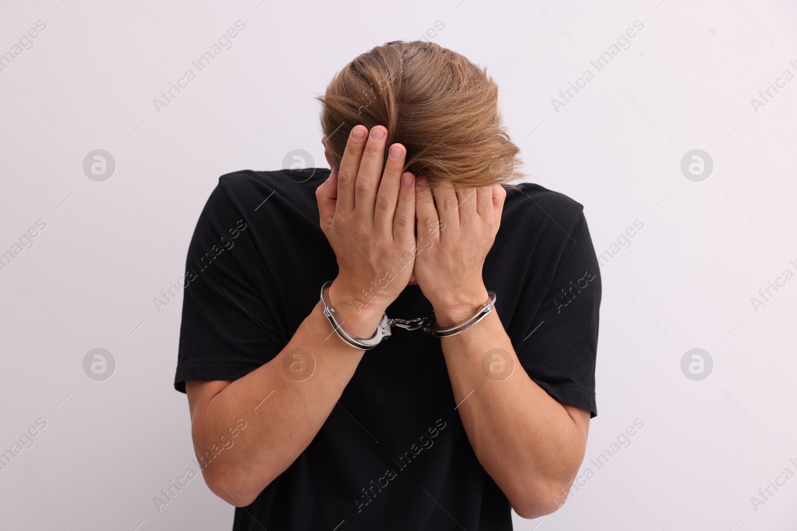 Man in metal handcuffs covering face with hands near light grey wall Photo of Man in metal handcuffs covering face with hands near light grey wall