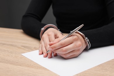 Photo of Woman in metal handcuffs writing something at wooden table against grey background, closeup