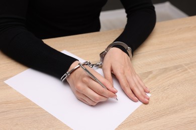 Photo of Woman in metal handcuffs writing something at wooden table indoors, closeup