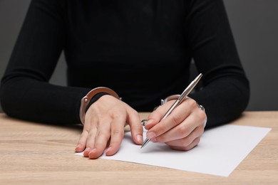 Photo of Woman in metal handcuffs writing something at wooden table against grey background, closeup