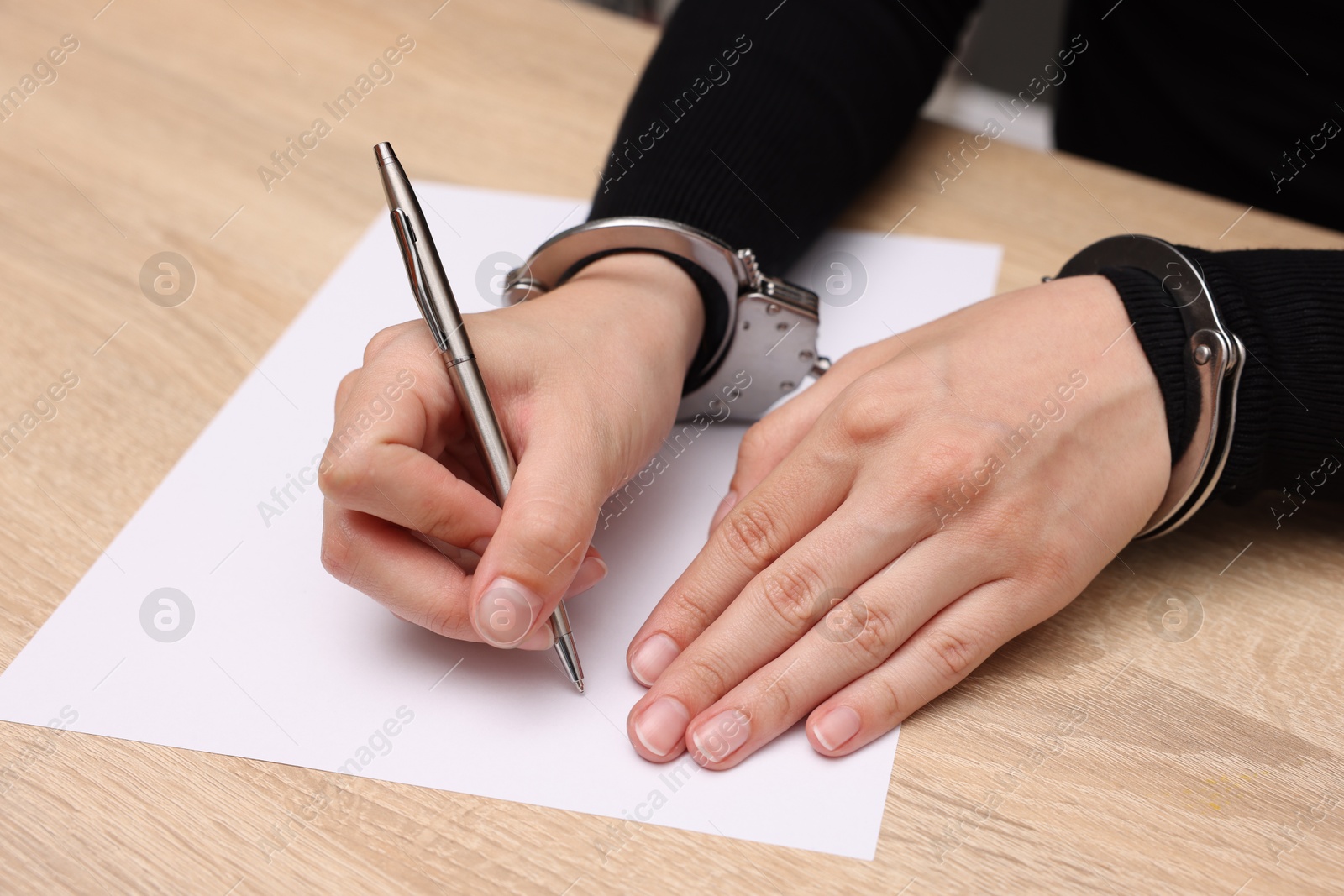 Photo of Woman in metal handcuffs writing something at wooden table indoors, closeup