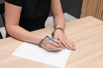Photo of Woman in metal handcuffs writing something at wooden table indoors, closeup