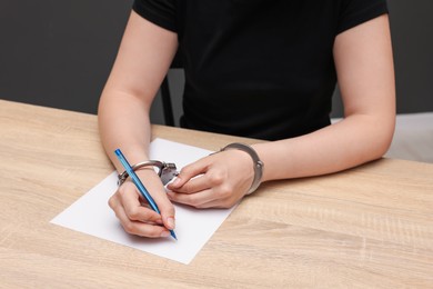 Photo of Woman in metal handcuffs writing something at wooden table against grey background, closeup