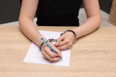 Photo of Woman in metal handcuffs writing something at wooden table indoors, closeup
