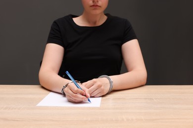 Photo of Woman in metal handcuffs writing something at wooden table against grey background, closeup