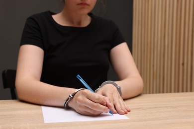 Photo of Woman in metal handcuffs writing something at wooden table indoors, closeup