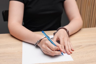 Photo of Woman in metal handcuffs writing something at wooden table indoors, closeup