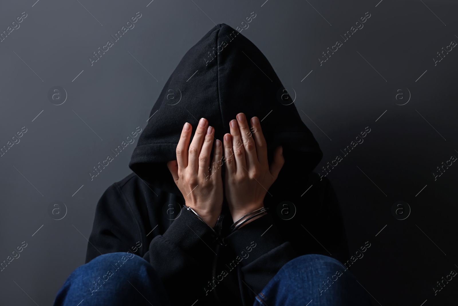 Photo of Woman in metal handcuffs and hoodie covering face with hands near grey wall