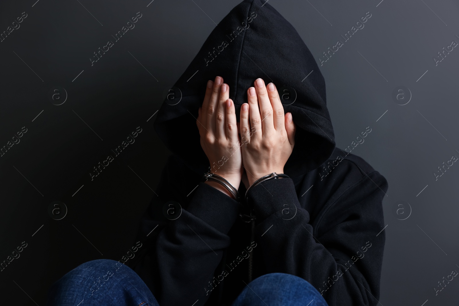 Photo of Woman in metal handcuffs and hoodie covering face with hands near grey wall