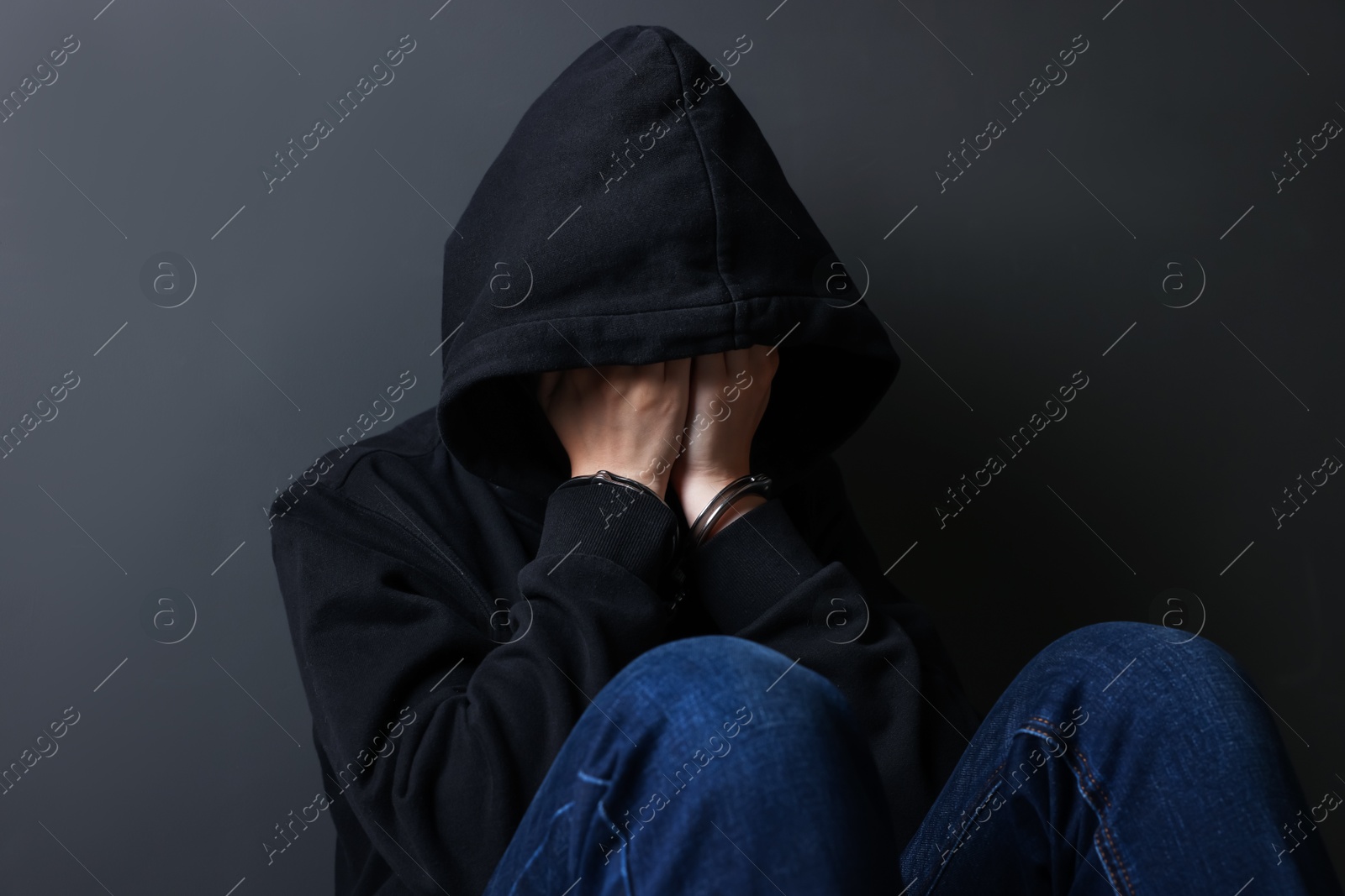 Photo of Woman in metal handcuffs and hoodie covering face with hands near grey wall
