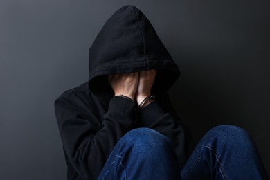 Photo of Woman in metal handcuffs and hoodie covering face with hands near grey wall