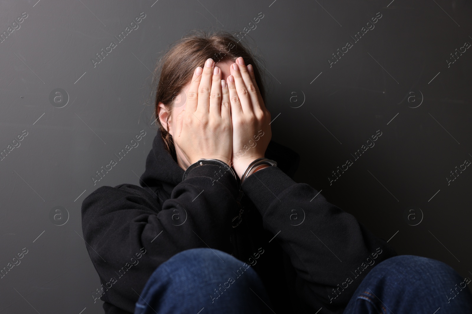 Photo of Woman in metal handcuffs and hoodie covering face with hands near grey wall