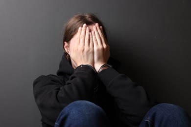 Photo of Woman in metal handcuffs and hoodie covering face with hands near grey wall