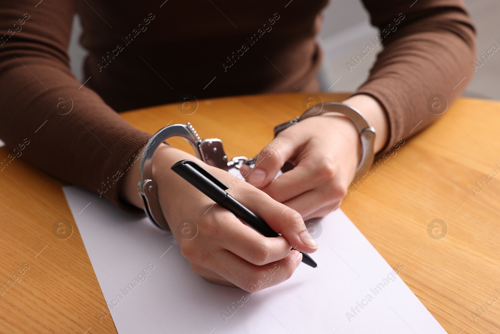 Photo of Woman in metal handcuffs writing something at wooden table against grey background, closeup