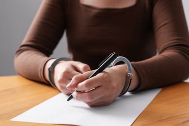 Photo of Woman in metal handcuffs writing something at wooden table against grey background, closeup