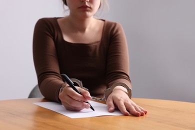 Photo of Woman in metal handcuffs writing something at wooden table against grey background, closeup