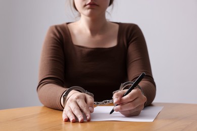 Photo of Woman in metal handcuffs writing something at wooden table against grey background, closeup
