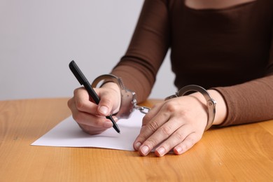 Photo of Woman in metal handcuffs writing something at wooden table against grey background, closeup