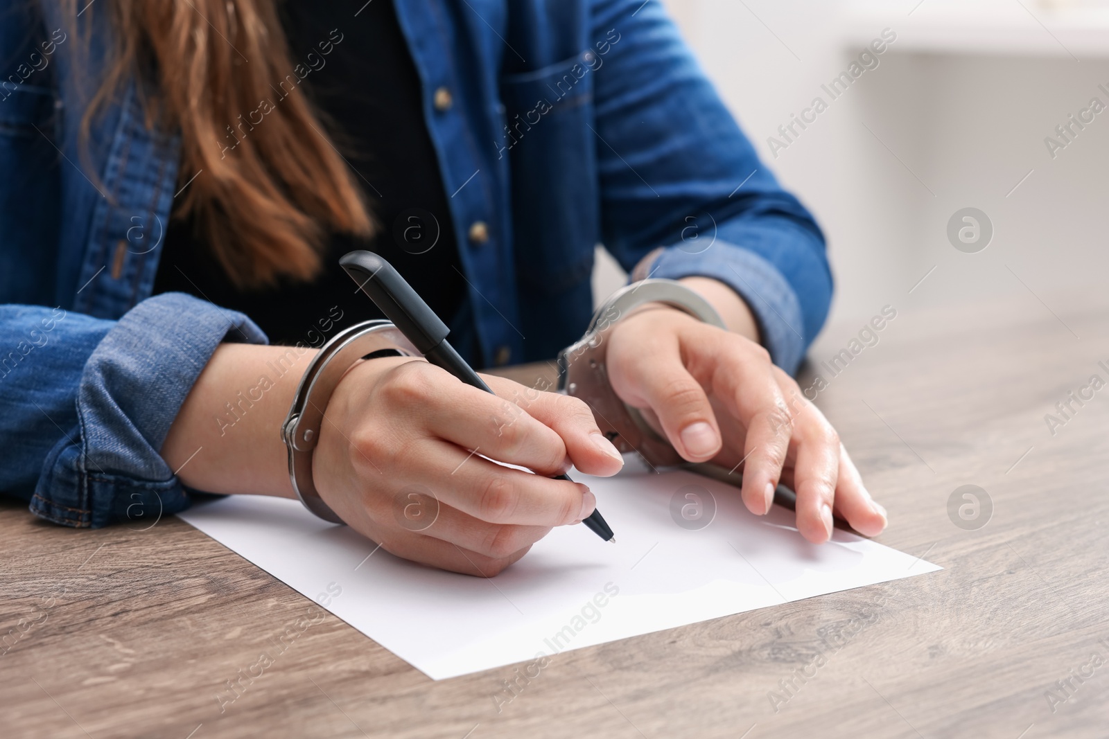 Photo of Woman in metal handcuffs writing something at wooden table indoors, closeup