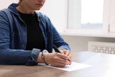 Photo of Woman in metal handcuffs writing something at wooden table indoors, closeup. Space for text