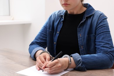 Photo of Woman in metal handcuffs writing something at wooden table indoors, closeup