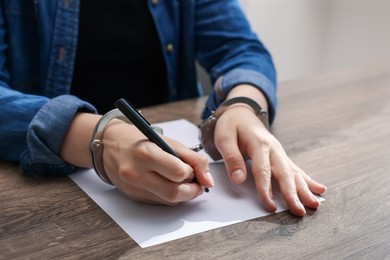 Photo of Woman in metal handcuffs writing something at wooden table indoors, closeup