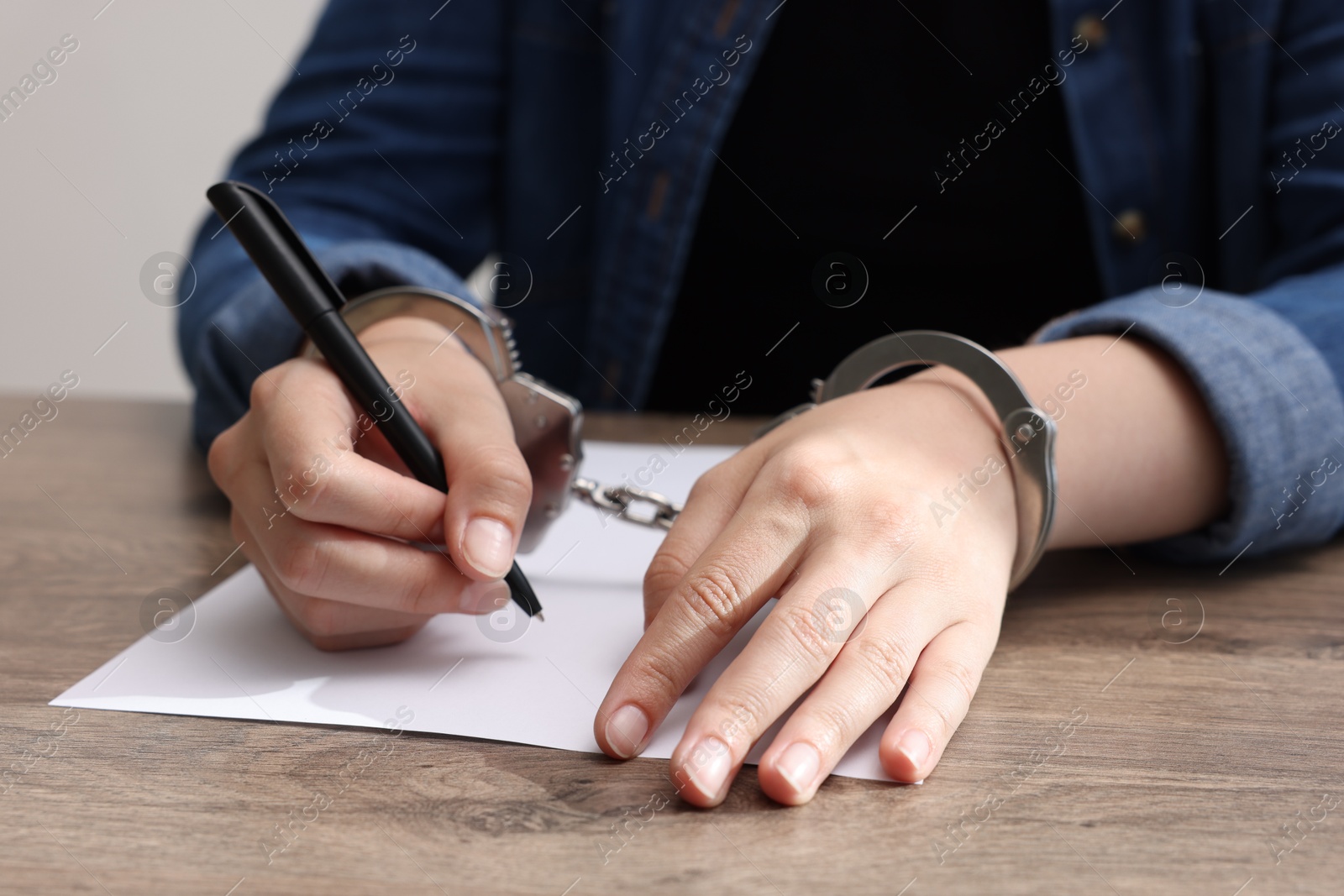 Photo of Woman in metal handcuffs writing something at wooden table indoors, closeup