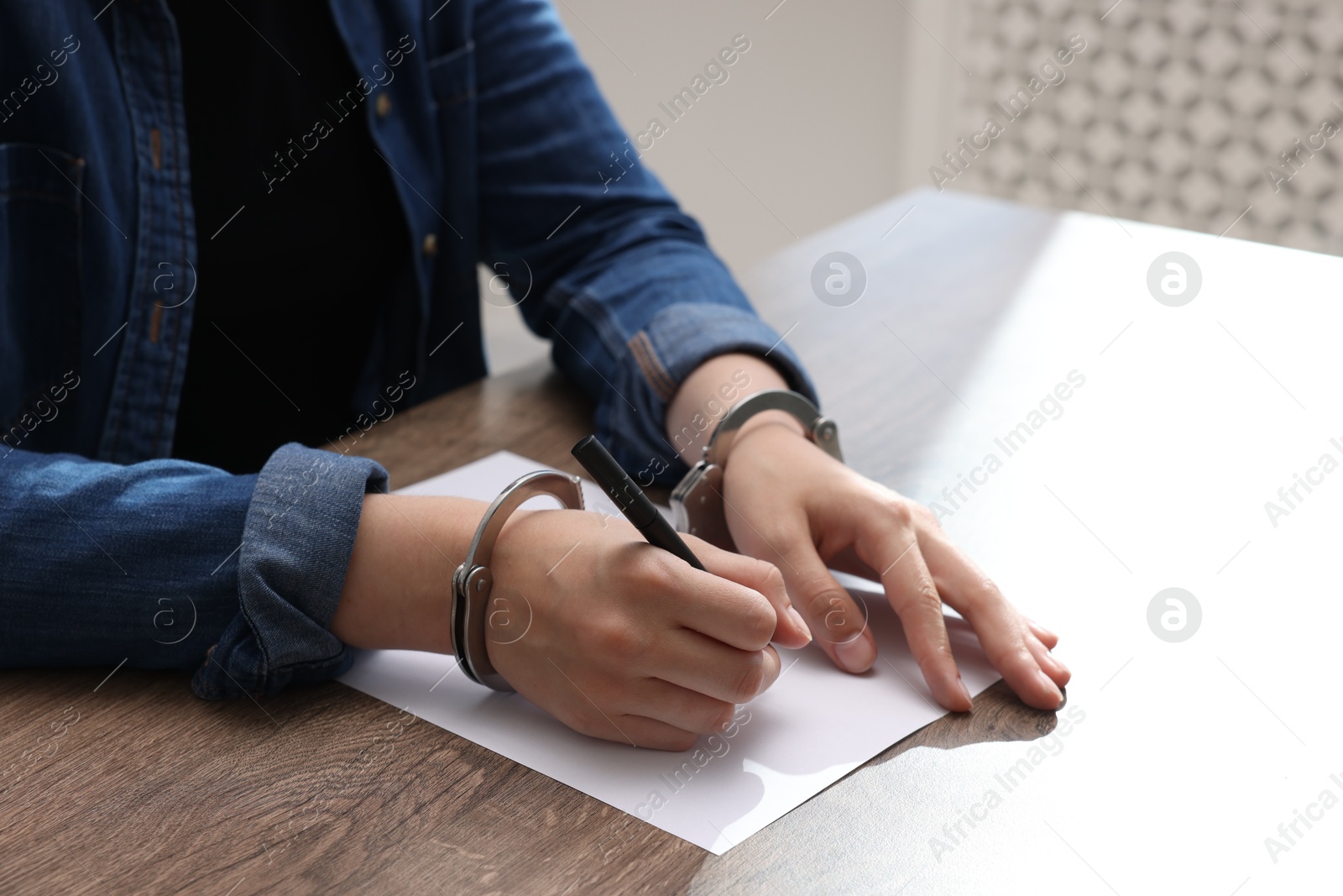 Photo of Woman in metal handcuffs writing something at wooden table indoors, closeup