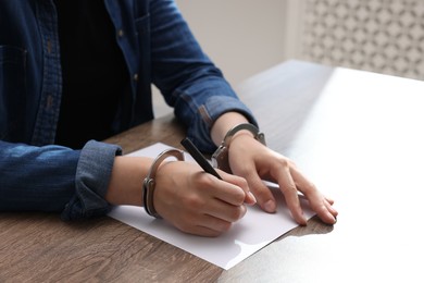 Photo of Woman in metal handcuffs writing something at wooden table indoors, closeup