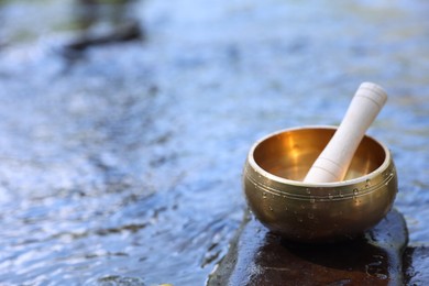 Photo of Tibetan singing bowl with mallet on stone in river, closeup. Space for text