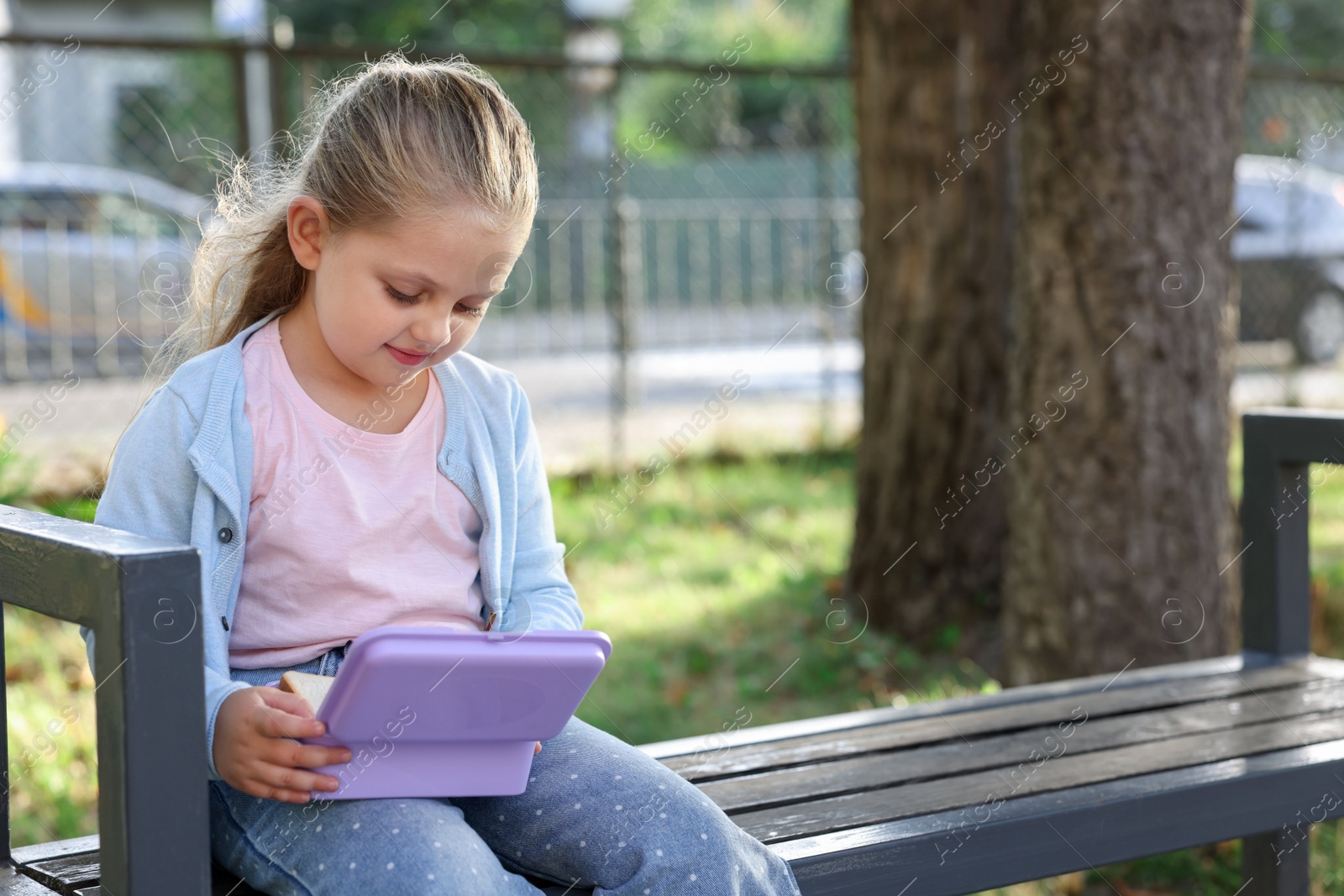 Cute little girl with school lunch box on bench outdoors, space for text Photo of Cute little girl with school lunch box on bench outdoors, space for text