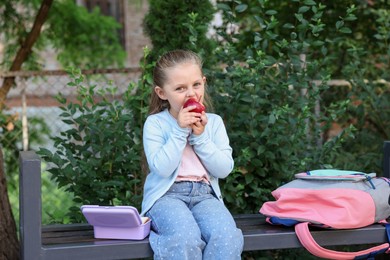 Photo of Cute little girl eating red apple on bench outdoors. School lunch
