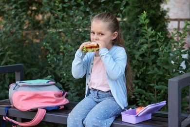 Photo of Cute little girl eating sandwich on bench outdoors. School lunch