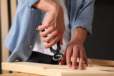 Photo of Man pulling metal nail out of wooden plank with pincers indoors, closeup