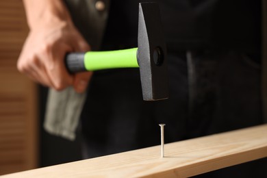 Photo of Man hammering nail into wooden plank indoors, closeup